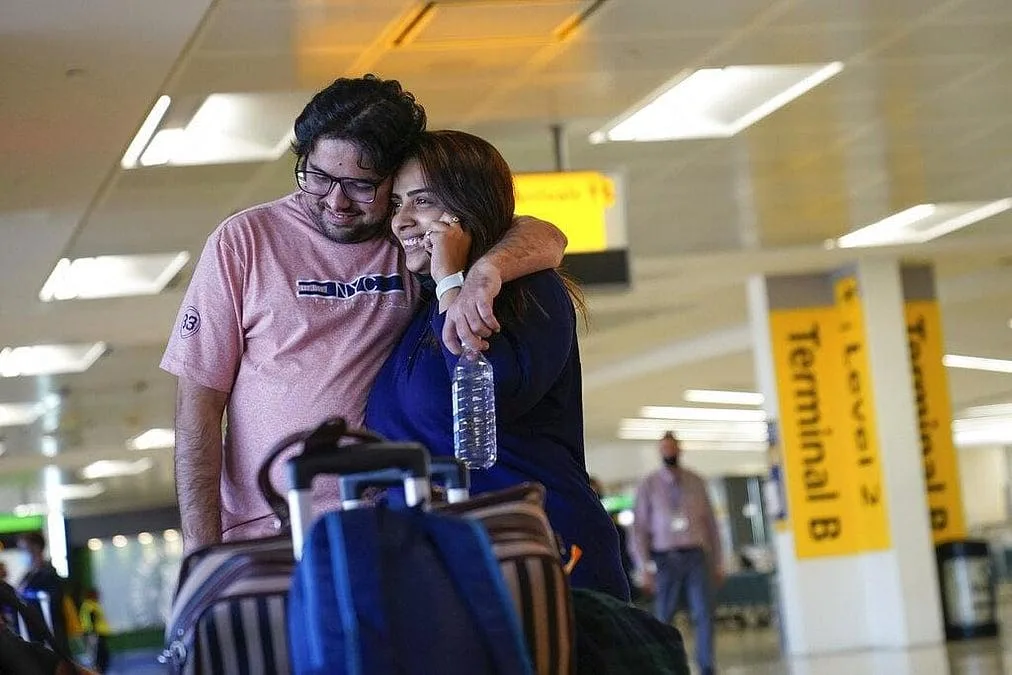 A young couple embrace with luggage in an airport terminal, smiling beneath overhead signs.