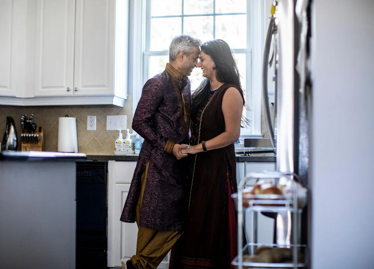 A couple in traditional Indian dress stand close together, foreheads touching and smiling, in a bright home kitchen.