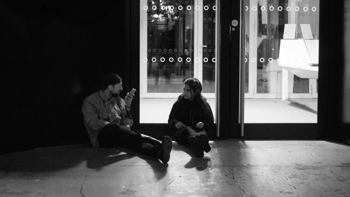A man and a woman sit on the ground facing one another at night, talking beside a lit glass doorway, in black and white.